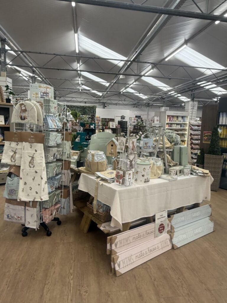 Indoor store display with home and garden gift items, including tea towels, signs, and decorative accessories on wooden tables.