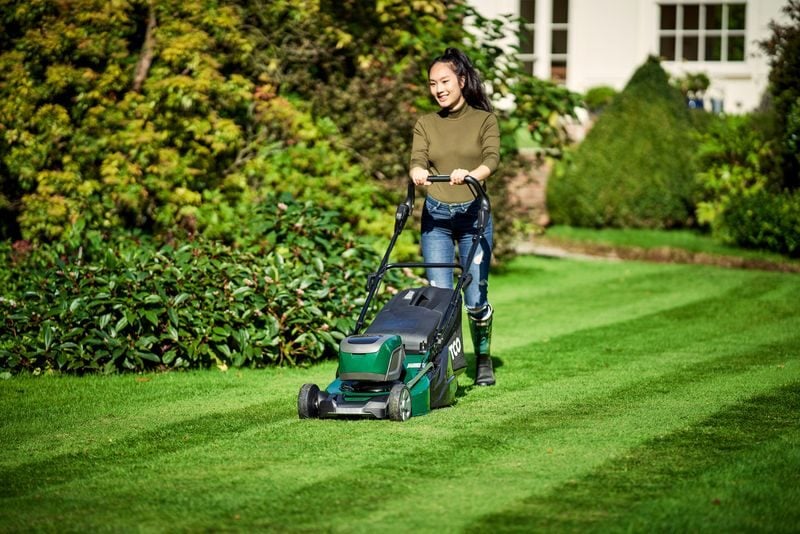 Woman mowing a freshly cut green lawn with a cordless electric lawn mower in a garden.