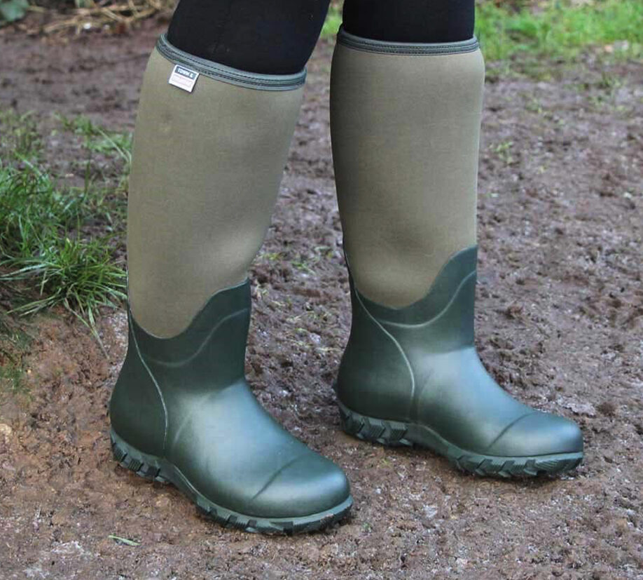Close-up of a person wearing green neoprene Wellington boots standing on muddy ground.