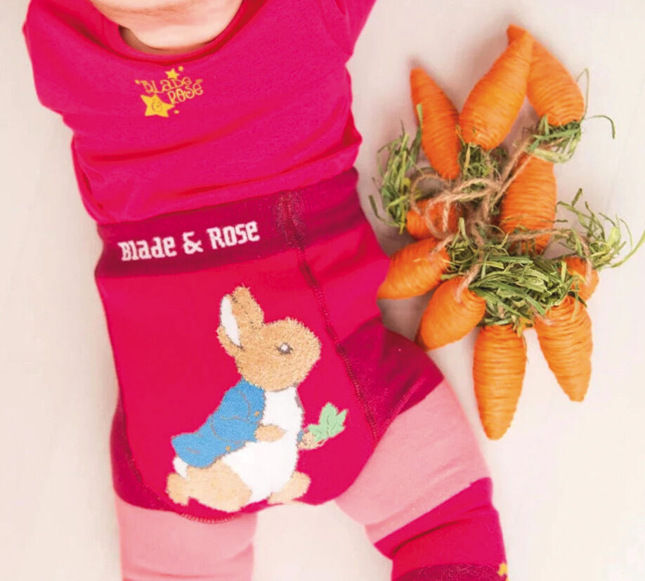 Baby wearing bright pink Blade & Rose leggings with a Peter Rabbit design, lying next to a bunch of toy carrots.