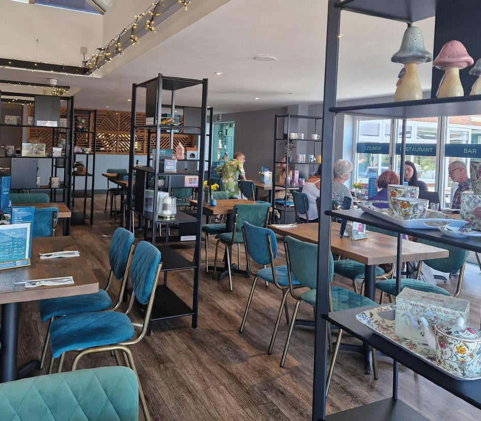 Daytime view of The Greenhouse Restaurant with teal chairs, wooden tables, and customers dining in a bright modern setting.