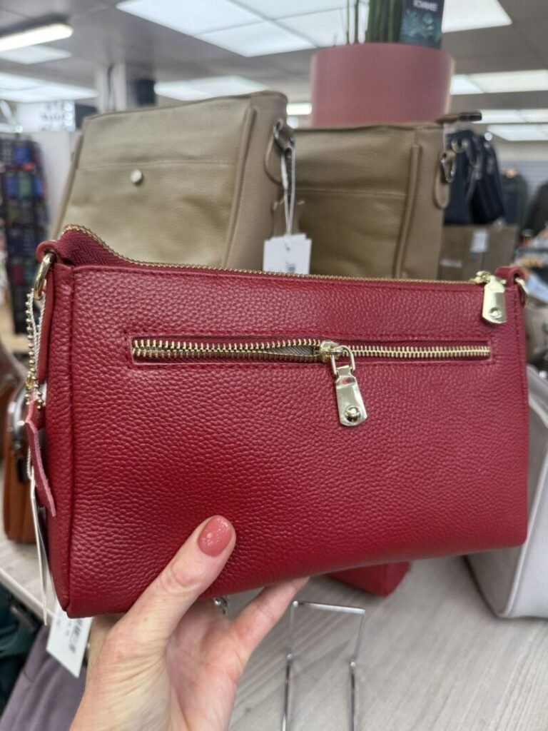 Person holding a red leather handbag with gold zippers in a shop display featuring various bags in the background.